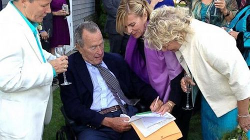 身穿西装的老布什坐在轮椅上，为一对同性恋新人签字证婚。